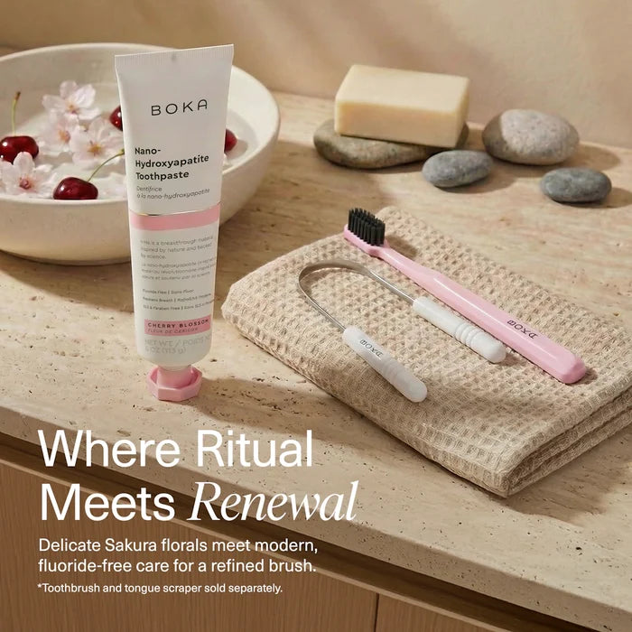 Boka toothpaste tube with a pink toothbrush and tongue scraper on a wooden surface with cherry blossoms.