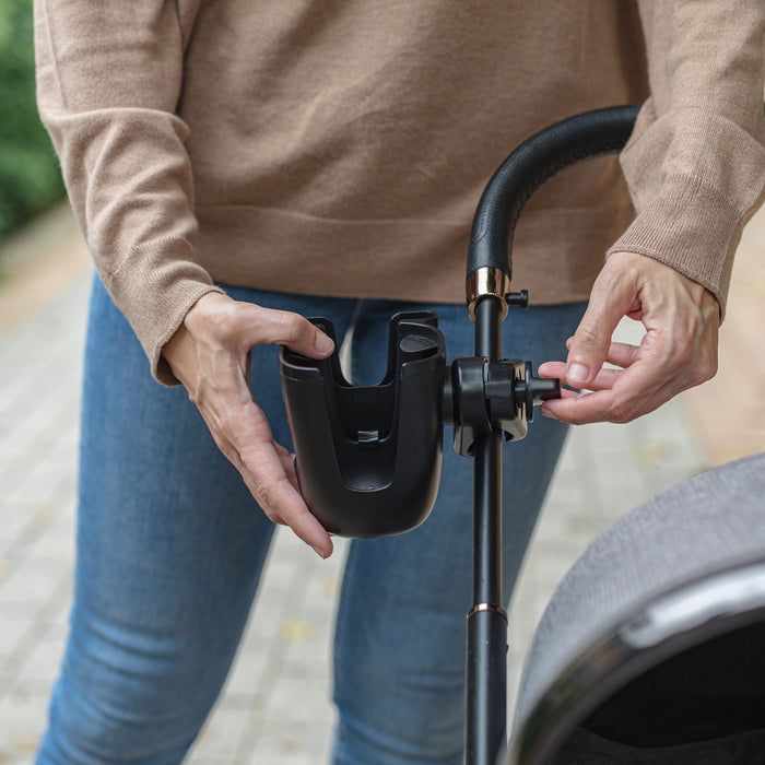 A person in a brown sweater and jeans attaches the Nuby Cup & Phone Holder to the handlebar of a baby stroller outdoors.
