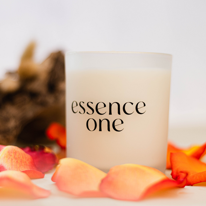 Close-up of a lit Essential Oil Soy Candle | Essence One in a frosted glass container surrounded by soft-focus rose petals, evoking a calming wellness atmosphere.