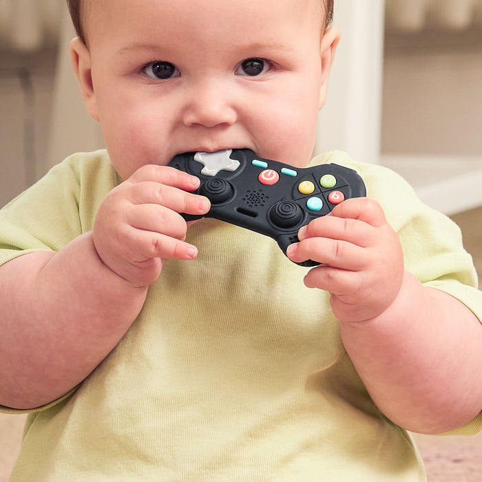 A baby wearing a light yellow shirt is sitting indoors and holding the Game Controller Teether with both hands, chewing on it.