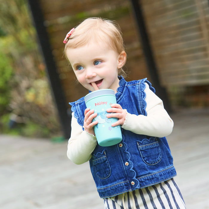 A young blonde-haired child in a blue denim vest and striped pants smiles outdoors while drinking from a Thirsty Kids Flexstraw Stainless Steel Cup | Whales.