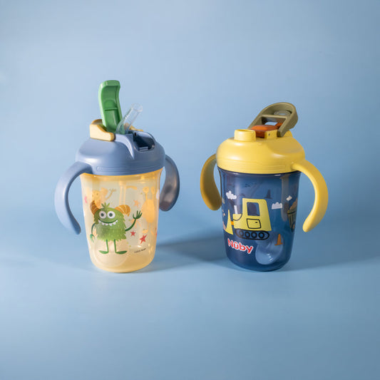 Two Nuby 2-in-1 Toddler Cups (Tractors & Monsters, 2 Pack) with handles rest on a light blue background. One is yellow with monsters, the other dark blue with a tractor. Each features a flip-top lid and weighted straw for easy sipping.