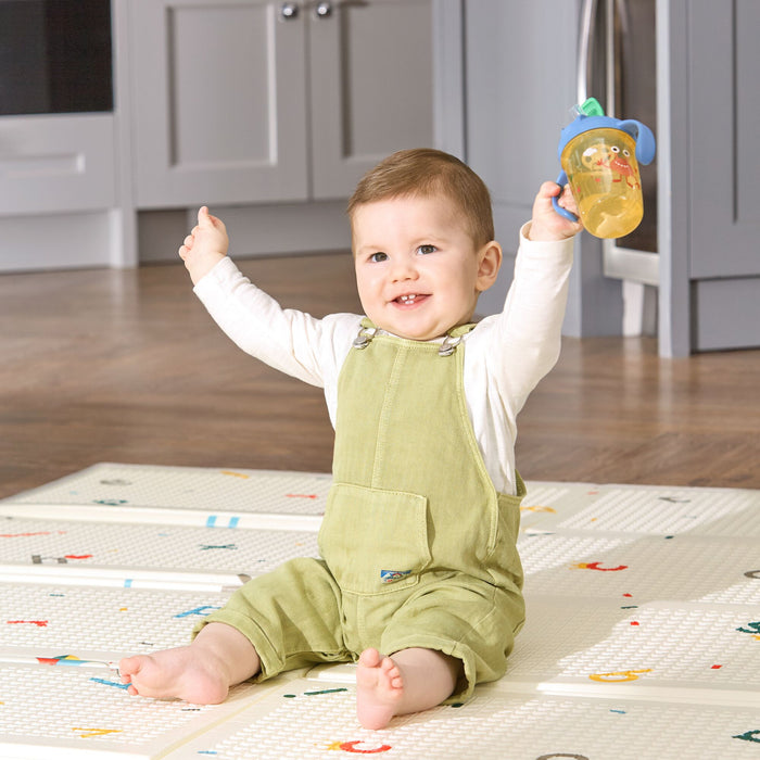 A happy baby in green overalls and a white shirt sits on a play mat in a bright kitchen, raising arms and holding the 2-in-1 Toddler Cup (2 Pack) | Tractors & Monsters with yellow liquid.