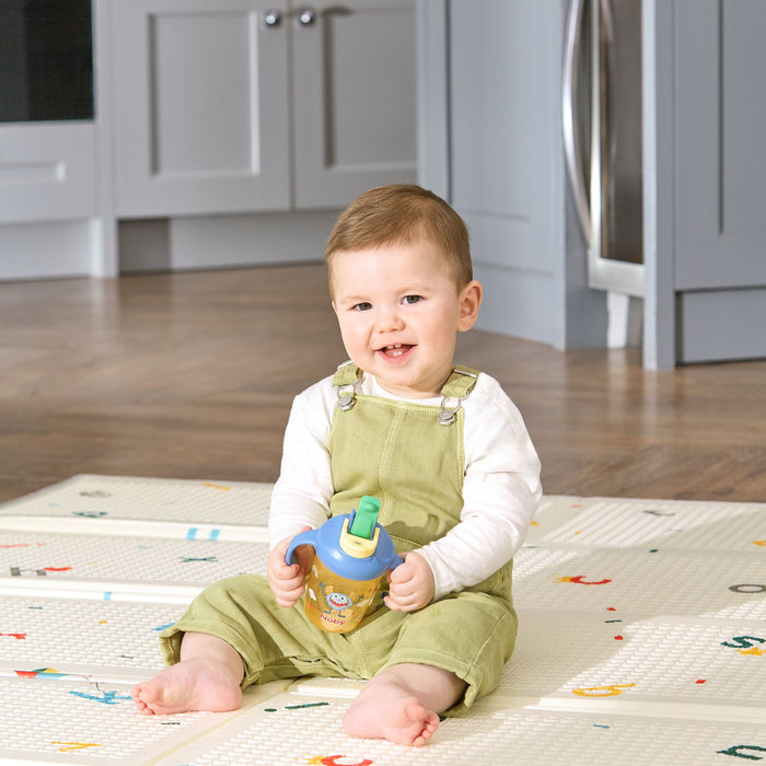 A smiling baby in light green overalls and a white shirt sits on a play mat, holding the 2-in-1 Toddler Cup (2 Pack) | Tractors & Monsters, with a kitchen in the background.