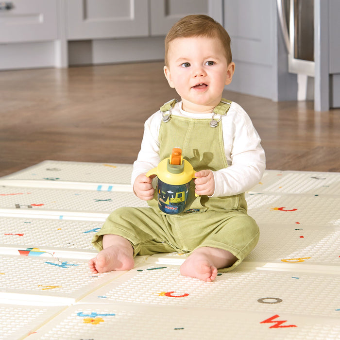 A baby in light green overalls and a white shirt sits barefoot on a patterned play mat, holding the 2-in-1 Toddler Cup (2 Pack) | Tractors & Monsters, with a kitchen visible in the background.