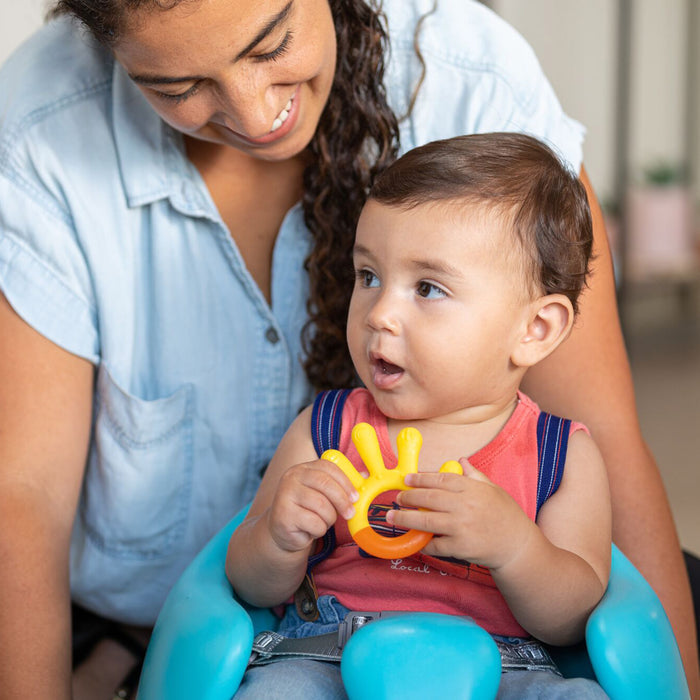 A smiling woman looks at a baby sitting in a blue seat, happily holding the 3 Step Baby Teether Set’s yellow hand-shaped silicone teether. The baby gazes up with an open mouth, and both appear happy and engaged.