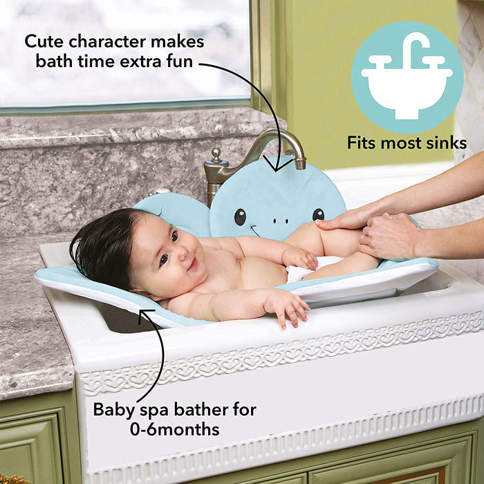 A smiling baby rests on the Baby Bath Sink & Tub Cushion, shaped like a plush blue whale, in a sink tub while an adult washes the baby’s arm. Text highlights: Cute design adds fun, fits most sinks, ideal for ages 0–6 months.