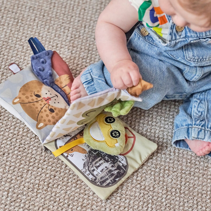 A baby in denim overalls sits on the floor, using one hand to turn a page of the Baby Sensory Book | City Bus—an engaging Montessori activity book with colorful city-themed illustrations and textures.