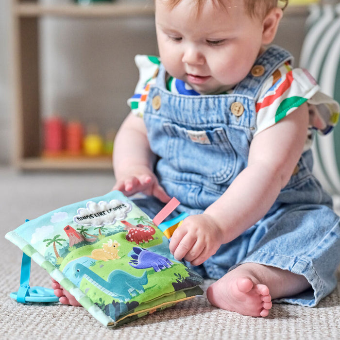 A baby in denim overalls sits on the floor, exploring the Baby Sensory Book | Dino—a colorful Montessori book with dinosaur illustrations and ribbon tags, ideal for sensory play or tummy time.