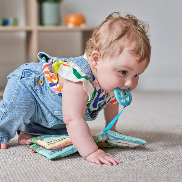 A baby in colorful clothes crawls on a carpet, holding a blue toy, with the Baby Sensory Book | Dino under their hands and a shelf with toys in the background.