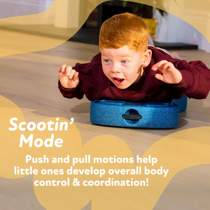 A young child with red hair lies on the beige Boogie Buggy 3-in-1 scooter board, using his hands to glide across a wooden floor. Text reads: Scootin Mode—push and pull actions help kids build body control & coordination!.