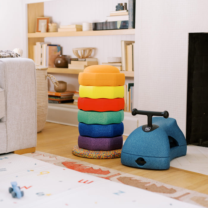A stack of colorful, rounded EPP foam cushions arranged in a rainbow sits beside the Boogie Buggy | Blue scooter in a cozy living room with shelves, books, a gray couch, and a lettered play mat on the floor.