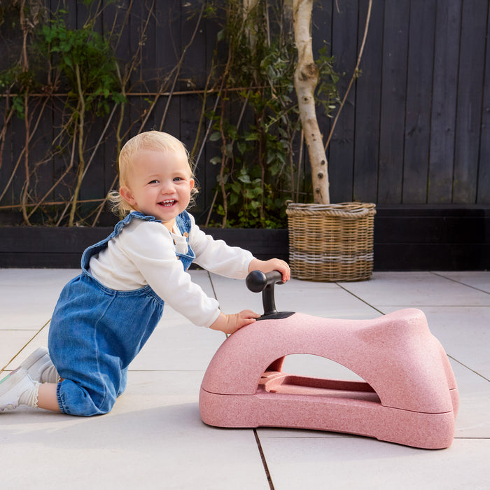 A smiling toddler in blue overalls kneels on a tiled patio, holding a Boogie Buggy | Pink 3-in-1 scooter shaped like an animal. A wicker basket and green plants sit against a dark wooden fence in the background.