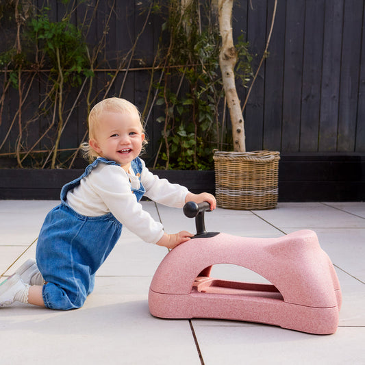 A smiling toddler in blue overalls kneels on a tiled patio, holding a Boogie Buggy | Pink 3-in-1 scooter shaped like an animal. A wicker basket and green plants sit against a dark wooden fence in the background.