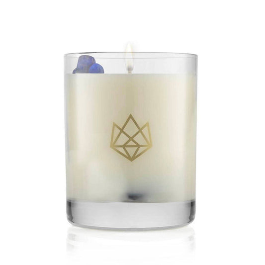 Lit white candle in a clear glass container with a subtle gold geometric logo, featuring small decorative blue stones, Calm Glass Candle | Aluminate Life.