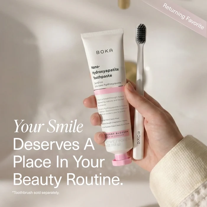 Hand holding a tube of Boka toothpaste with a toothbrush, against a neutral background.