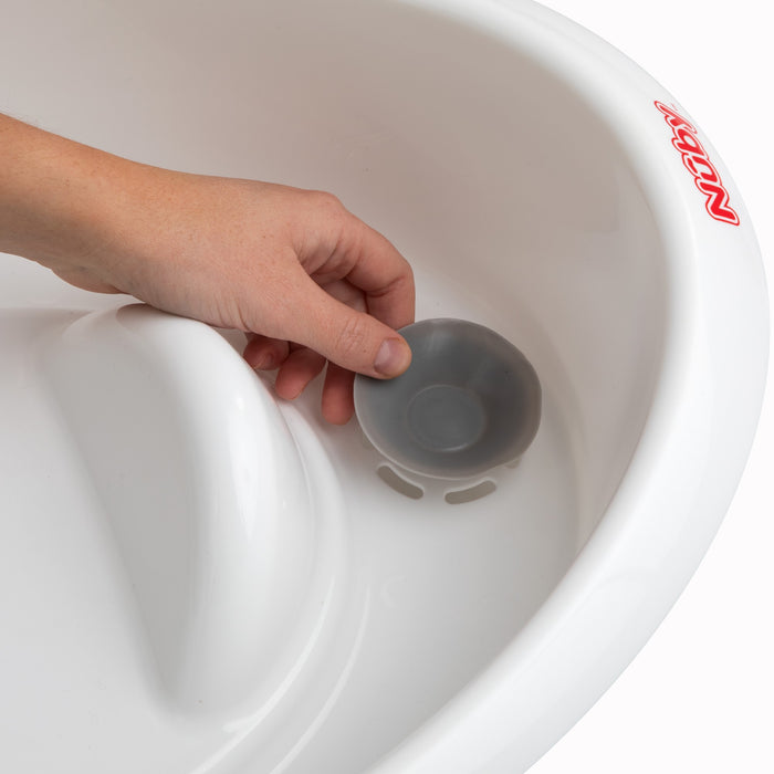 A hand places or removes a round gray drain plug from the Comfort Baby Bathtub, a white plastic newborn tub by Nuby, with the Nuby logo shown in red.