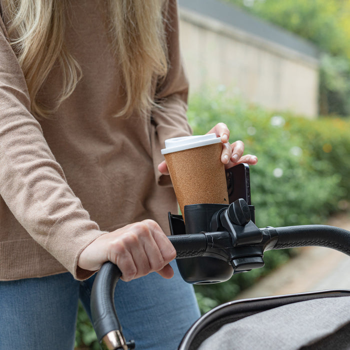 A person in a beige sweater and blue jeans pushes a stroller outdoors with greenery behind them. The Nuby Cup & Phone Holder is attached to the handle, securely holding their coffee cup as they walk.