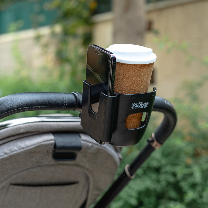 A Nuby Cup & Phone Holder, in black, attaches to a stroller handle and securely holds a brown coffee cup with a white lid and a smartphone, with blurred greenery and a wall visible in the background.