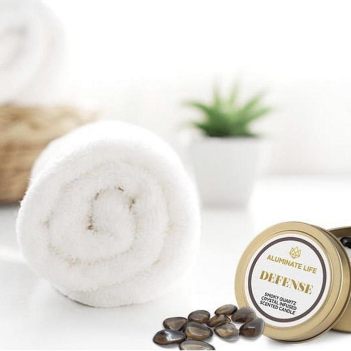 Defense Candle Tin | Aluminate Life displayed with smooth black stones and a rolled white towel, suggesting a soothing wellness and spa setting.