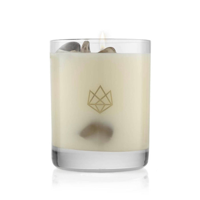 Lit creamy white candle with small decorative stones inside a clear glass holder featuring a subtle geometric logo, Defense Glass Candle | Aluminate Life.