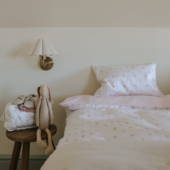 Bedroom with a bed featuring pink bedding and a small stool with a plush toy and bag against a wall-mounted lamp.