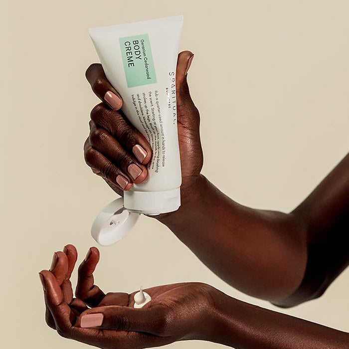 A person with dark skin applying Geranium Cedarwood BODY CRÉME | Sparitual, dispensing a small amount of cream onto their palm for skincare and body care purposes.