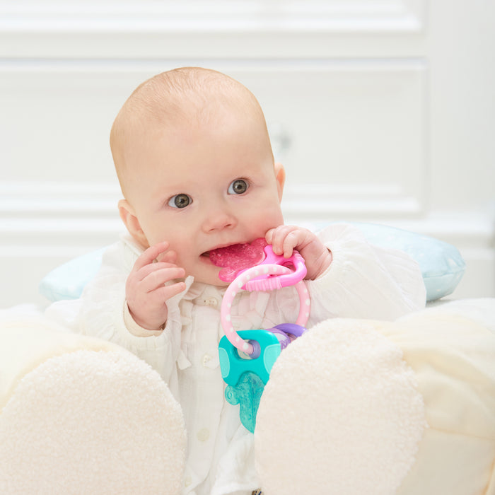 A baby in white clothing sits on a soft chair, chewing the pink part of the IcyBite Teether Keys | Pastels by Nuby, with a white dresser in the background.