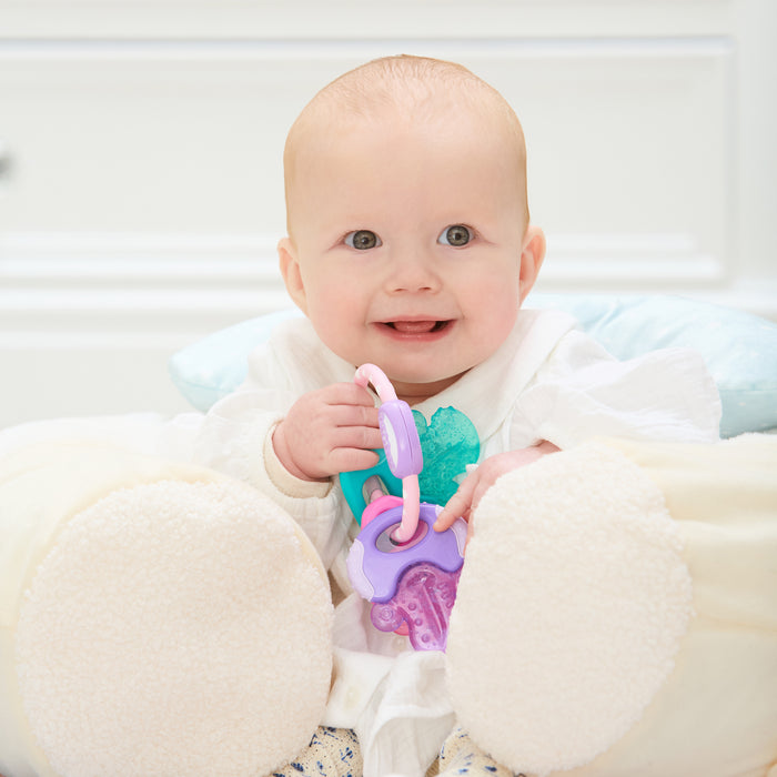 A smiling baby sits propped up with a soft pillow, wearing a white outfit and holding colorful Nuby IcyBite Teether Keys | Pastels.