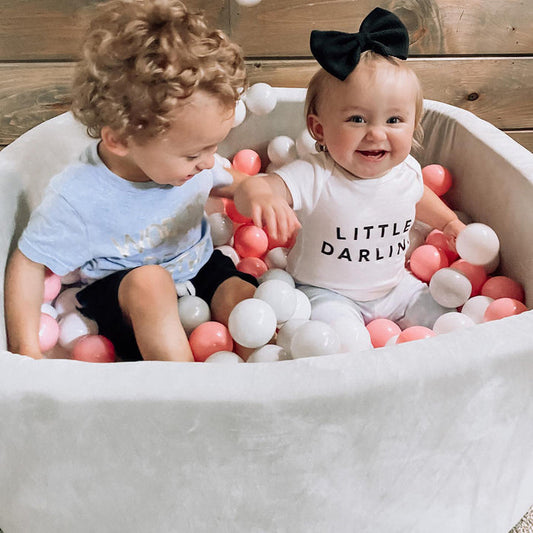 Two young children sit in the Kids' Ball Pit for Home with 200 Balls in pink. The girl, wearing a big black bow and Little Darling shirt, and the boy in a light blue shirt enjoy playing among the colorful balls.