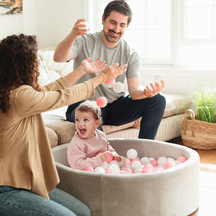 A young child smiles in a Kids' Ball Pit for Home with 200 Balls | Pink, surrounded by white and pink balls, while two adults joyfully toss balls in the air inside a bright, cozy living room.