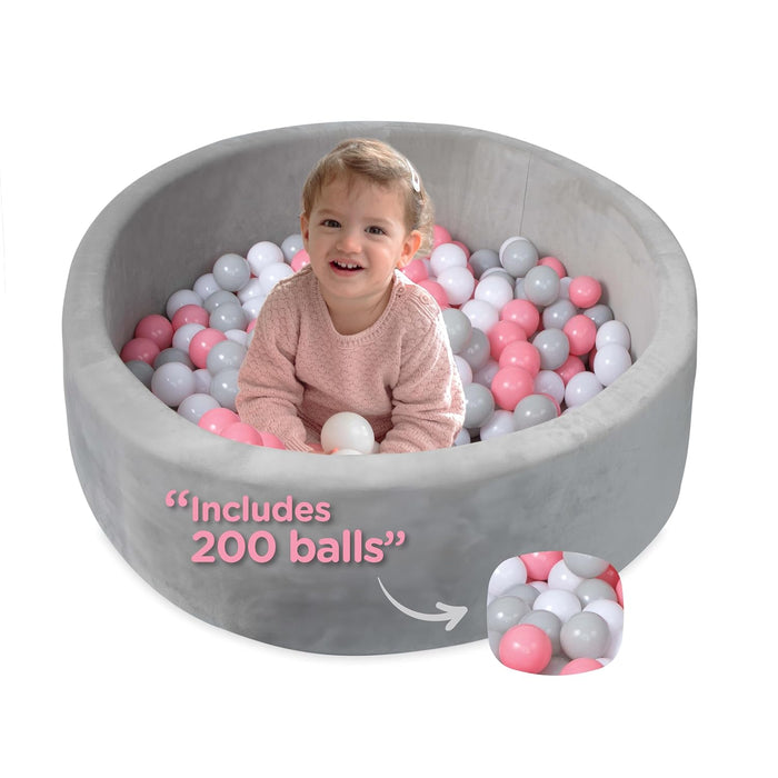 A smiling toddler plays in the Kids' Ball Pit for Home with 200 Balls | Pink, surrounded by pink, white, and gray balls. Text on the image reads "Includes 200 balls," with a close-up inset of the colorful balls.