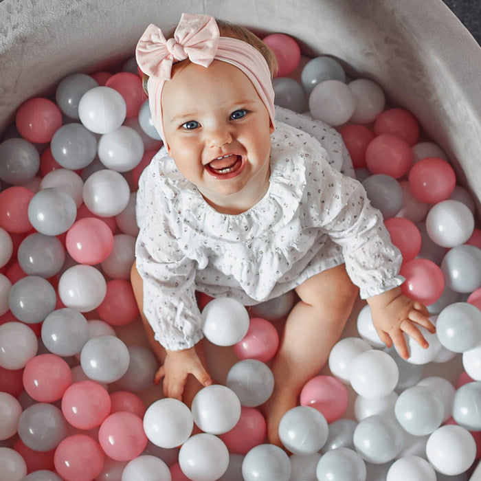 A baby with blue eyes and a pink bow headband smiles while sitting in the Kids' Ball Pit for Home with 200 Balls | Pink, filled with pink, white, and gray balls and wearing a white long-sleeve outfit.