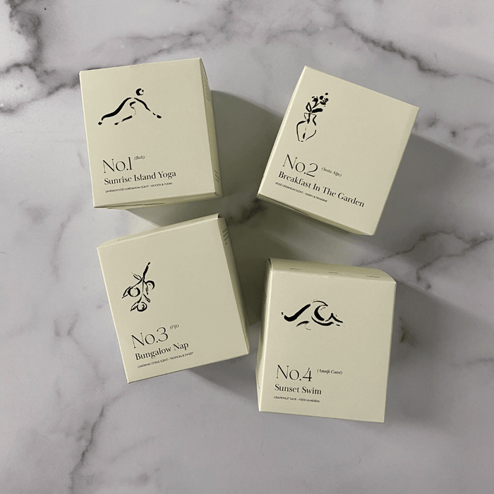 Four minimalist cream-colored candle boxes with simple black illustrations and text, featuring No. 4 Sunset Swim (Amalfi Coast) Candle | Lucky Owl among others, arranged on a marble surface.