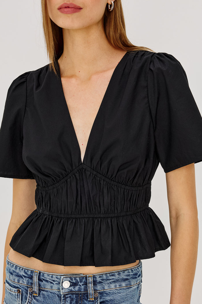 Orla Shirt in Cotton Poplin With Short Puff Sleeves, Deep V-Neckline, Shirred Shoulders, Elastic Waist and Underbust, and Peplum Hem in Black Colorway - Upper Body Front View Featured on Model