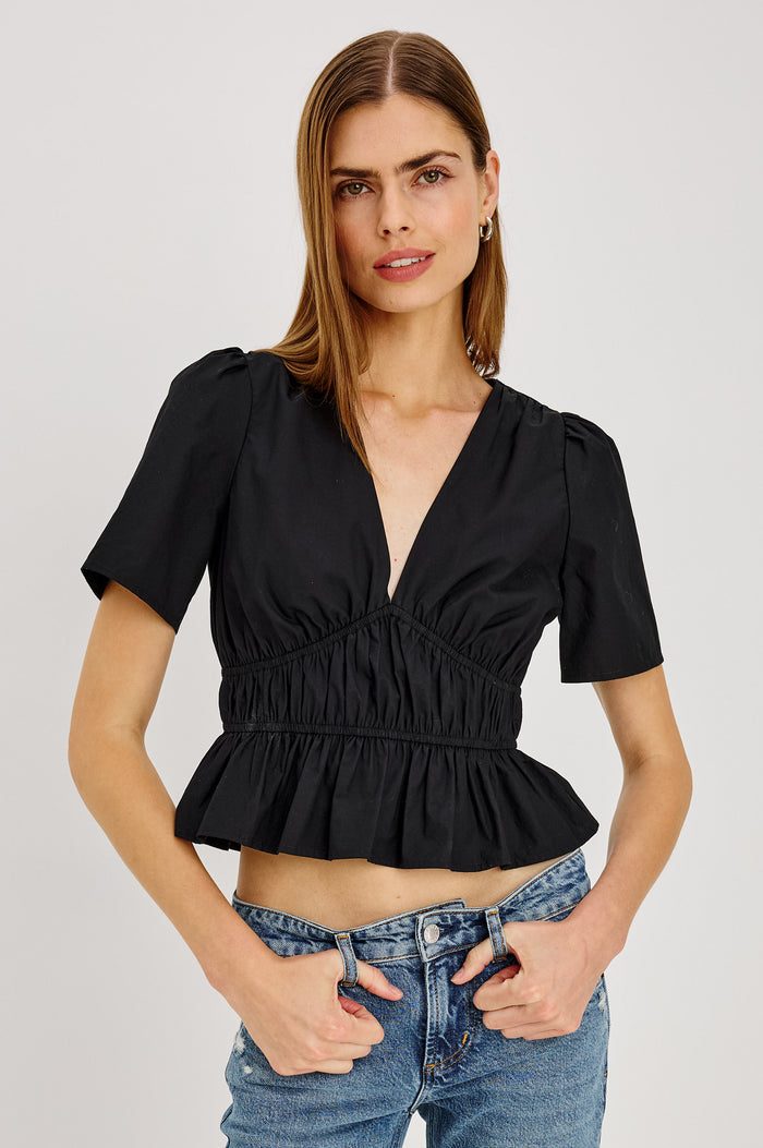 Orla Shirt in Cotton Poplin With Short Puff Sleeves, Deep V-Neckline, Shirred Shoulders, Elastic Waist and Underbust, and Peplum Hem in Black Colorway - Upper Body Front View Featured on Model