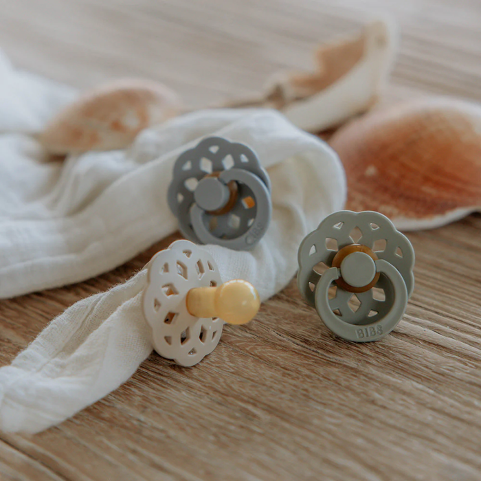 Three baby pacifiers on a wooden surface with a white cloth and shells in the background.