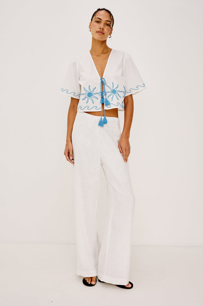Pari Short-Sleeve Cropped Top with V Neck, Front Tie Closures, and Sun Embroidery Details in Blue Stitching Against White Fabric - Full Body Front View Featured on Model