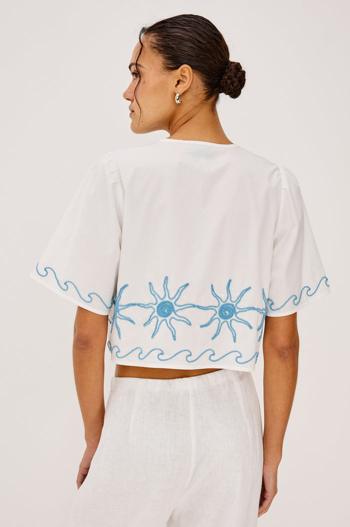 Pari Short-Sleeve Cropped Top with V Neck, Front Tie Closures, and Sun Embroidery Details in Blue Stitching Against White Fabric - Upper Body Back View Featured on Model