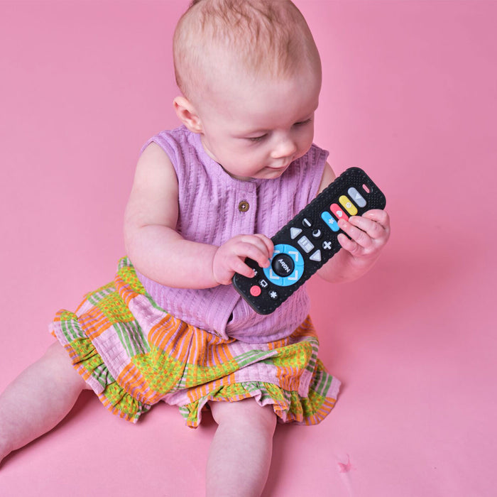 A baby in a purple top and colorful skirt sits on a pink surface, looking down and holding the Press Play Bundle with both hands.