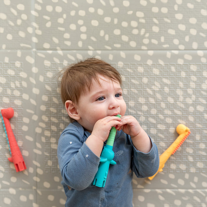 A baby in blue sits on a patterned mat, holding and chewing a silicone teether from the Pretend Play Teething Bundle by Nuby US, with red and yellow toys from the same set nearby for soothing teething relief.