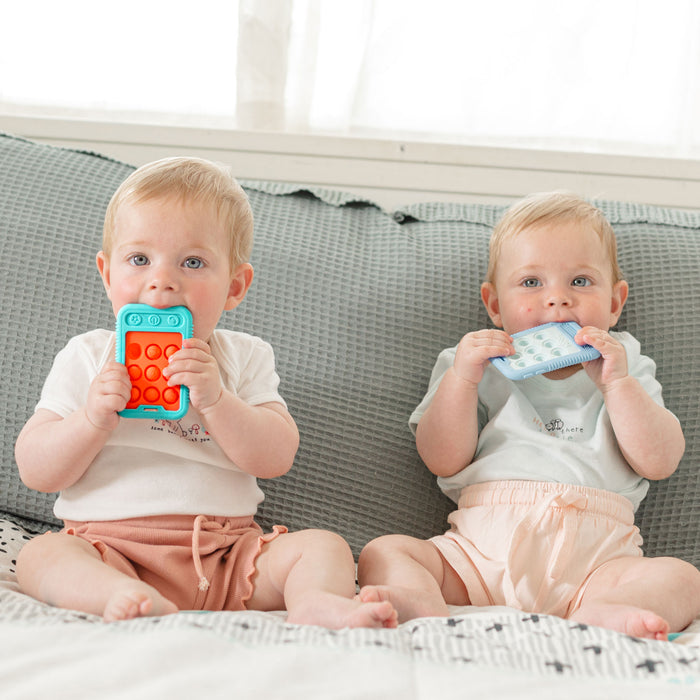 Two light-haired babies sit on a sofa, each chewing on a colorful Giggle Bytes Sensory Popper Teething Toy. Both wear light shirts and peach shorts, with a soft blanket and window providing a gentle background.