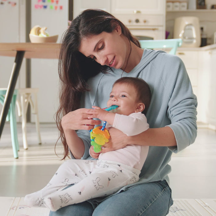 In a bright, modern kitchen, a woman holds a smiling baby in her lap as the baby chews on the Nuby US Pretend Play Teething Bundle for soothing relief. Both look relaxed and happy.