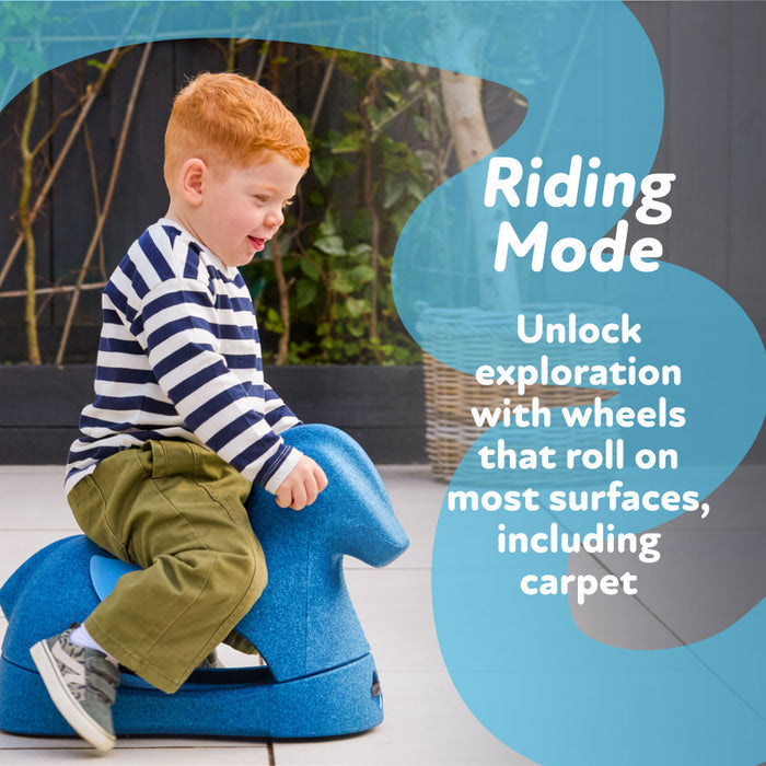 A young child with red hair, in a striped shirt and green pants, rides the Rock 'N Ride | Pink indoors. Text reads: Riding Mode. Unlock exploration with wheels designed to roll smoothly across most surfaces, even carpet.