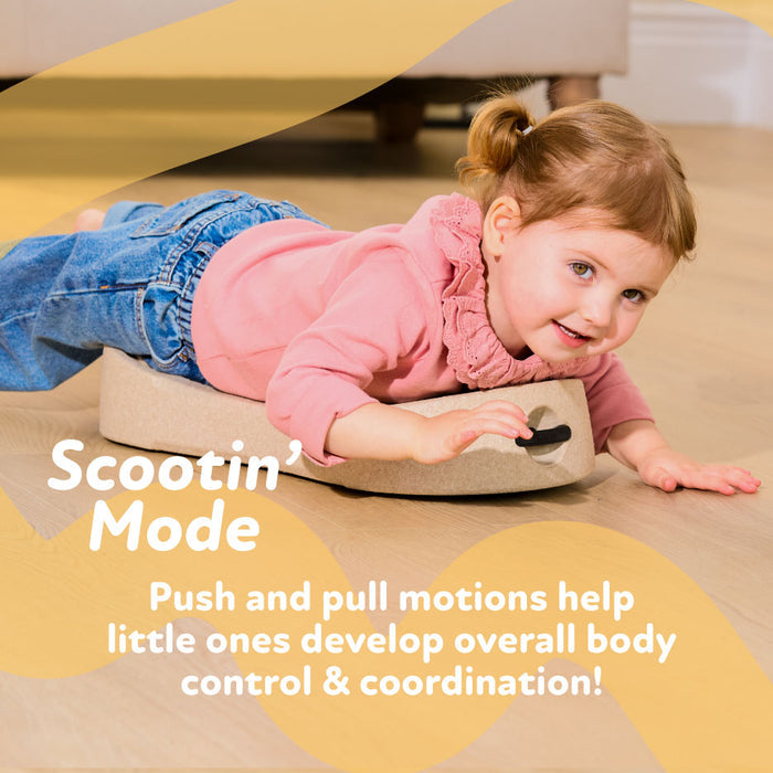 A young child smiles while lying on their stomach on the Rock 'N Ride | Sage toddler scooter, pushing themselves forward. Text reads: Scootin Mode—push and pull motions help develop body control & coordination!.