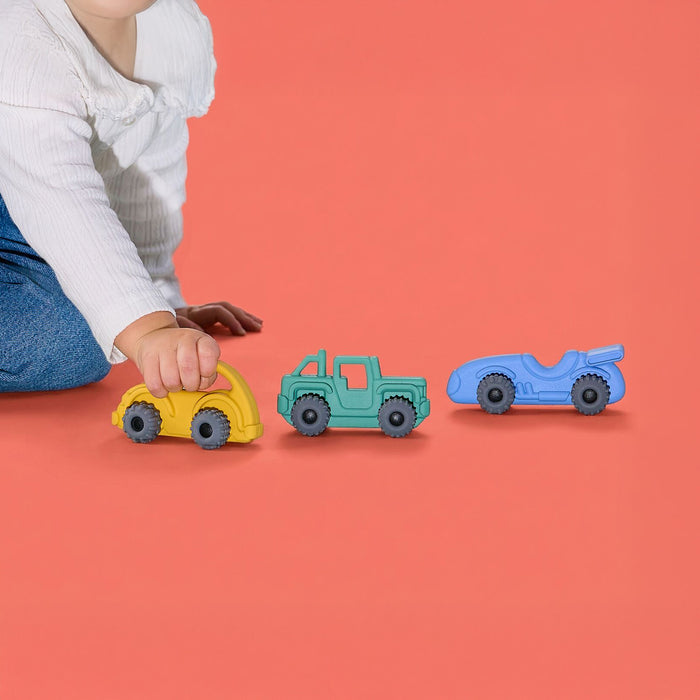 A child in a white shirt and blue jeans plays with three colorful toy vehicles—a yellow car, a green truck, and a blue race car—on a vibrant coral background.