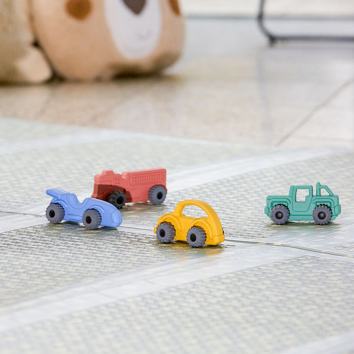 A Nuby Silicone Vehicle Teether Toy | Car sits near four colorful toy vehicles—a blue motorcycle, red fire truck, yellow car, and green jeep—on a patterned play mat, with a plush bear partially visible in the background.