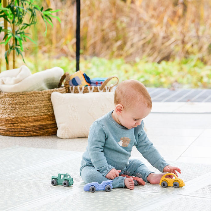 A baby in a blue outfit sits on the floor with three toy cars—green, blue, and yellow—and the Nuby Silicone Vehicle Teether Toy | Fire Engine. A wicker basket, pillow, and greenery with an outdoor view are behind them.