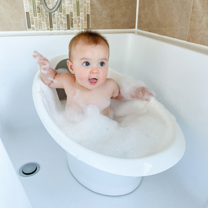 A baby sits in a small white bathtub filled with bubbles, looking surprised and playful with one hand raised. The cozy tub is placed inside a larger bathtub in a tiled bathroom, creating an adorable bath time scene.