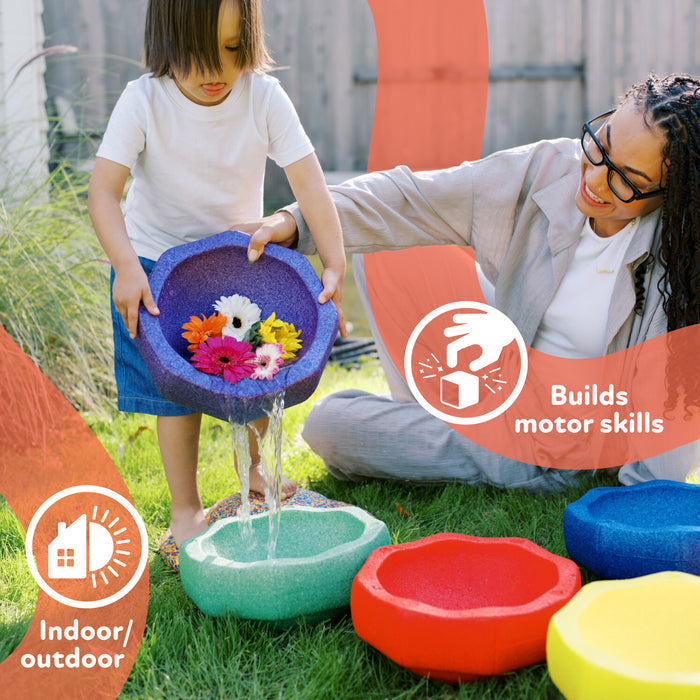 A child pours water and flowers from a purple bowl into a blue one outdoors, as an adult smiles nearby. StepPebbles and FREE Balance Board | Pastel bowls on the grass are perfect for play or pairing with foam stepping stones to boost motor skills.